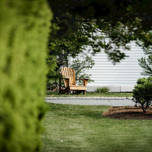 A wooden chair sits in a shaded, grassy yard near a white building, framed by trees and shrubs in the foreground, creating a serene scene.