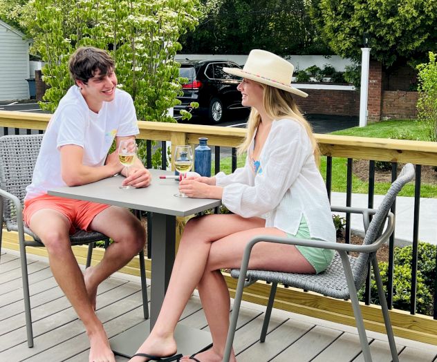 Two people are sitting on a deck at a small table outdoors, enjoying drinks and smiling at each other. It appears to be a sunny day.