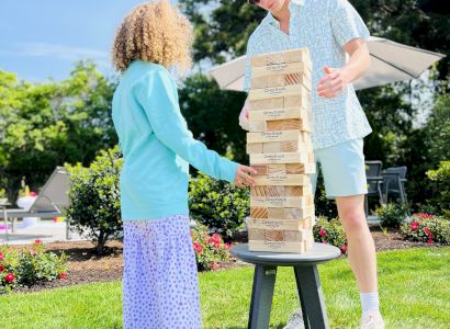 Two people are playing a game of giant Jenga on a stool outdoors in a garden, with a sunny sky and trees in the background.