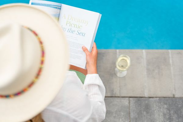 A person wearing a hat reads a book by a poolside with a glass of white wine beside them. The book's title page reads 